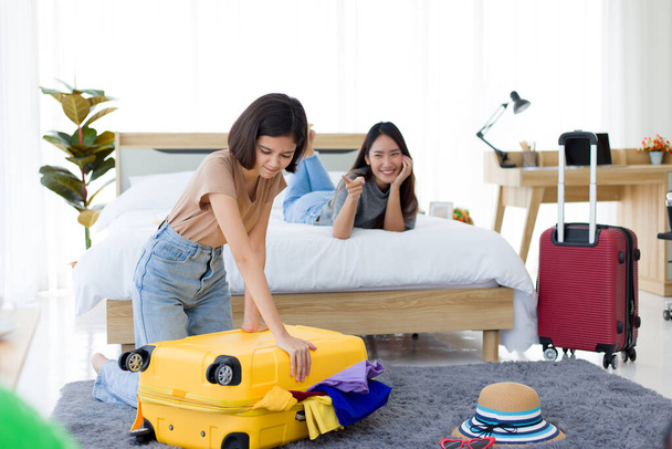 Young Asian woman preparation travel suitcase at home while her friend lying on the bed and were laughing at him Because of too many clothes. - 写真・画像