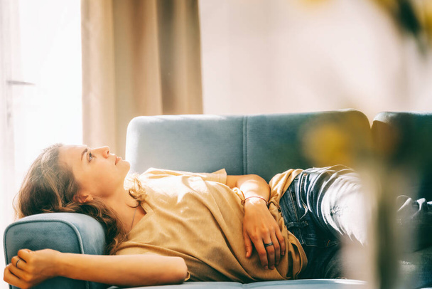 Jeune belle femme bouclée allongée à la maison sur un canapé bleu et relaxante, portrait de profil - Photo, image