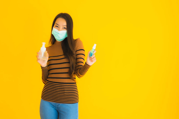 Portrét krásná mladá asijská žena s maskou alkohol sprej a gel láhev v ruce na ochranu před koronavirem nebo covid19 na žlutém izolovaném pozadí - Fotografie, Obrázek