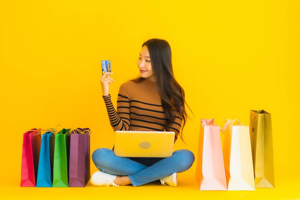 Hermoso retrato joven asiática mujer sentarse en el suelo con el ordenador portátil o el uso de la tarjeta de crédito para las compras en línea alrededor de la bolsa de compras en fondo amarillo
 - Foto, imagen
