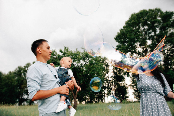 glückliche junge Eltern und Sohn haben Spaß beim Spielen mit Seifenblasen auf der Wiese an einem Sommertag - Foto, Bild