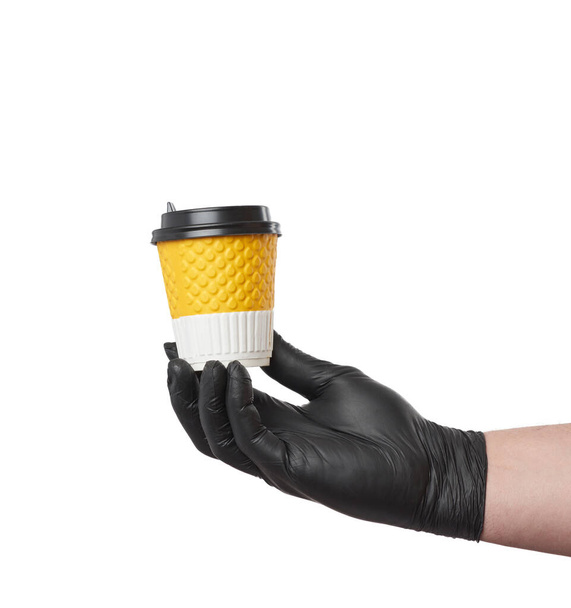 χέρι σε ένα μαύρο γάντι λατέξ κρατά ένα κύπελλο μιας χρήσης από χαρτόνι και ένα πλαστικό καπάκι σε λευκό φόντο, ασφαλή και ανέπαφη παράδοση on-line παραγγελιών κατά τη διάρκεια επιδημιών - Φωτογραφία, εικόνα