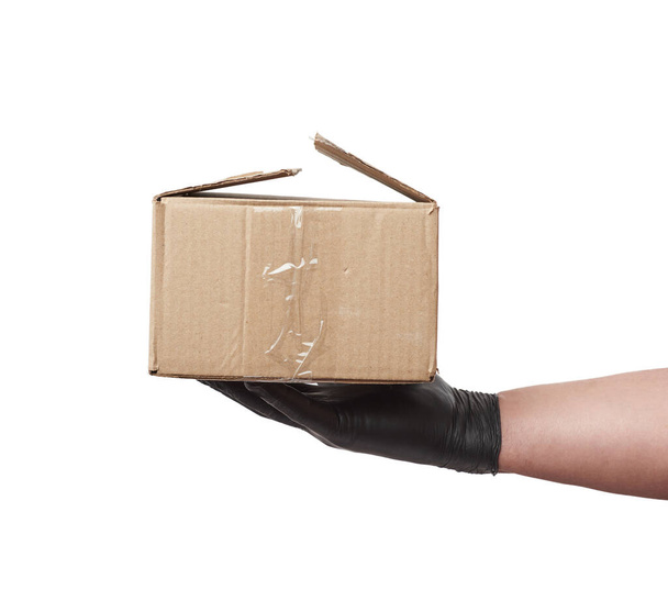 Ruka v černé latexové rukavici drží kartónovou krabici z hnědého papíru kraft na bílém pozadí, bezpečné a bezkontaktní doručení on-line objednávek během epidemie - Fotografie, Obrázek