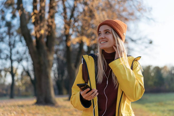 Kaunis hymyilevä nainen yllään keltainen sadetakki ja tyylikäs hipster hattu kuuntelee musiikkia katselee pois ulkona. Nuori tunteellinen turisti kävelee puistossa. Inspiraatio, matkakonsepti - Valokuva, kuva