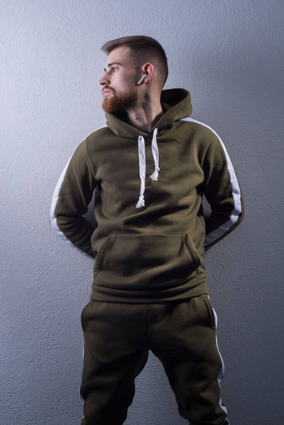 Studio portrait of a young bearded guy of twenty-five years old. in a sports jacket with a swamp-colored hood. On a gray background. Advertising photo of a handsome guy. in a sports warm jacket with a hood. green military color. In wireless headphone - Foto, Imagem