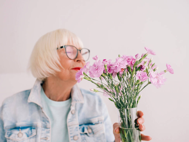 senior femme élégante en lunettes et jeans veste odeur oeillet fleurs dans un vase. Fleurs, retraite, profession, concept de passe-temps - Photo, image