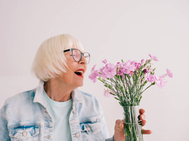anziana donna elegante in occhiali e jeans giacca odore fiori di garofano in vaso. Fiori, pensionamento, professione, hobby concept - Foto, immagini