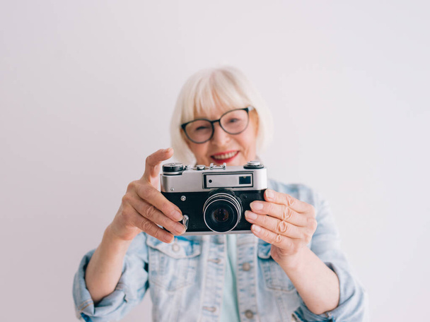 sênior (velho) mulher elegante com cabelo grisalho e em óculos e jeans jaqueta tirando fotos com câmera de filme. Idade, hobby, anti idade, vibrações positivas, conceito de fotografia
 - Foto, Imagem