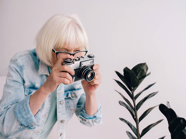 白髪のスタイリッシュなシニアやフィルムカメラで花の写真を撮るメガネやデニムジャケット。年齢、趣味、反年齢、肯定的な振動、写真の概念 - 写真・画像