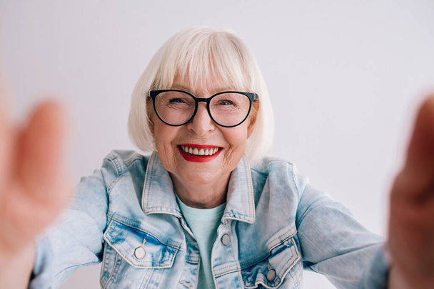 senior (old) stylish woman with gray hair and in glasses and jeans jacket hugging you or making selfie. Love, hugs, anti age, positive vibes concept - Photo, Image