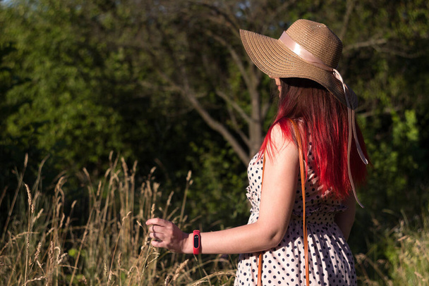 Uma jovem com cabelo vermelho está vestida com um vestido bege em bolinhas pretas e segura uma lâmina de grama, a menina está de pé em um campo entre a grama. Verão, fundo
 - Foto, Imagem