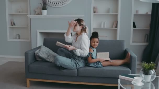 Portrait little african girl daughter back to back sits on sofa with her mother, foster parent or nanny in living room. Standing there holding books, looking at camera smiling. Concept of happy family - Filmagem, Vídeo