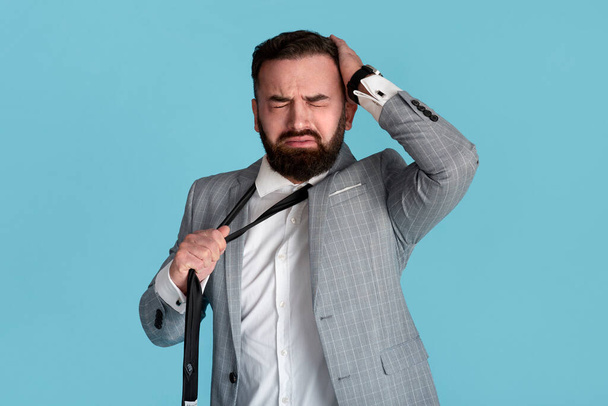 Desperate young office worker grasping head and pulling his tie over blue background - Photo, Image