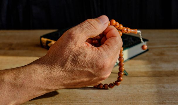 Hand of a senior Muslim man fingering a rosary made of stone on a background of a book of Quran. Close up Copy space - Foto, Imagen