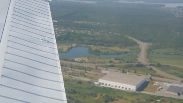 De witte vleugel van een passagiersvliegtuig tegen de top uitzicht op de natuur achtergrond van een prachtig blauw meer, vliegt over een vijver, groene bossen en kleine huizen en steden. Reisconcept - Video