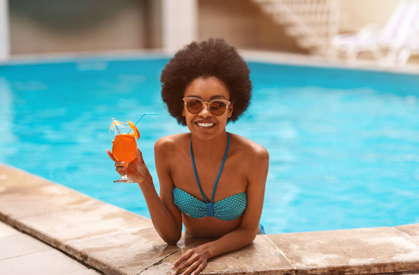 Portrait of carefree black woman in swimsuit with cocktail drink at pool on tropical resort - Foto, Imagem