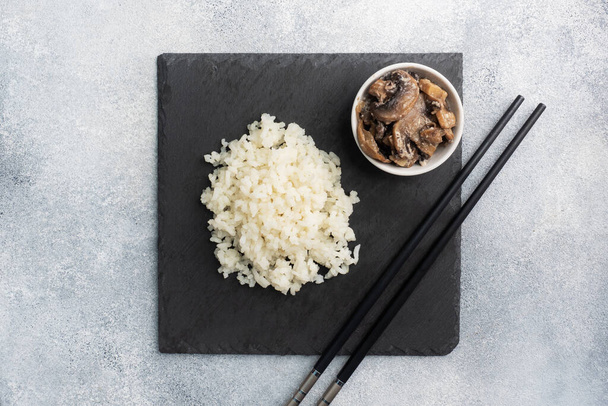 Boiled white rice and mushrooms in sour cream on a black stand with chopsticks. Grey concrete background Copy space. - Foto, immagini