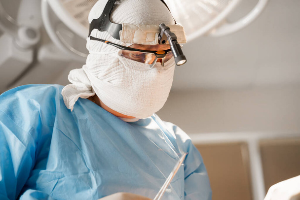 Хирург с фарой и ножницами делают пластическую операцию в медицинской клинике. Увеличение груди пластическая операция и коррекция в медицинской клинике
 - Фото, изображение