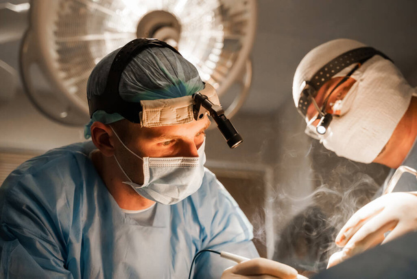 2 surgeons with headlamp do plastic operation in medical clinic. Chest augmentation plastic operation and correction in medical clinic. Group of professional medics in medical masks do plastic surgery. - Photo, Image