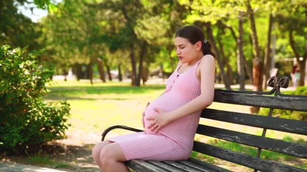 Une fille enceinte est assise sur un banc de parc, et son estomac commence à faire mal et elle commence à respirer correctement. femme enceinte caressant le ventre. - Séquence, vidéo