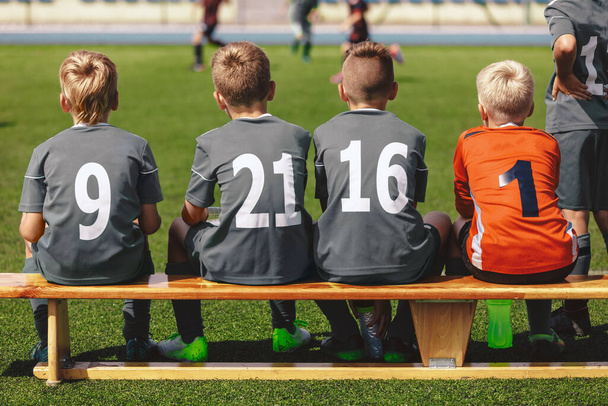 Futbol takımındaki çocuklar yedek kulübesinde oturuyorlar ve final turnuvasına hazırlar. Sırtında oyuncu numaraları olan spor üniformalı okul çocukları. Okulun Genç Seviye Takımında Futbolcular - Fotoğraf, Görsel