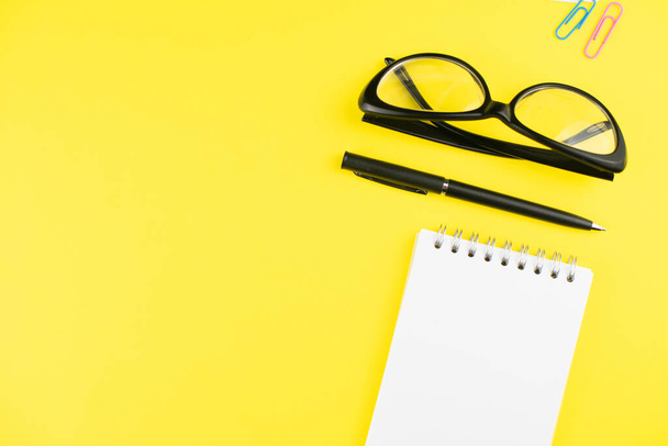 Black glasses, pen, notepad and colorful clips on yellow background. Flat lay. Copy space. Workplace in the office.  - Photo, Image