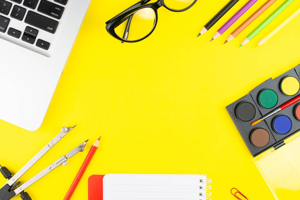 Notepad, veelkleurige potloden, verdeler, zwarte bril, kleurrijke clips, olieverf voor tekening en laptop op gele achtergrond. Bovenaanzicht met kopieerruimte.  - Foto, afbeelding