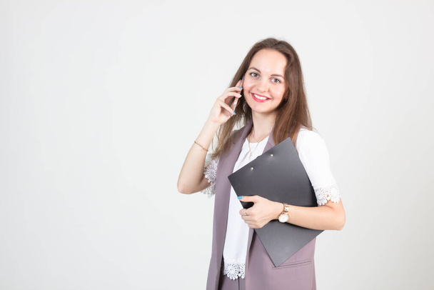 όμορφη νεαρή γυναίκα στις επιχειρήσεις αυστηρά ρούχα θέτει σε ένα λευκό τοίχο με ένα τηλέφωνο στα χέρια της - Φωτογραφία, εικόνα
