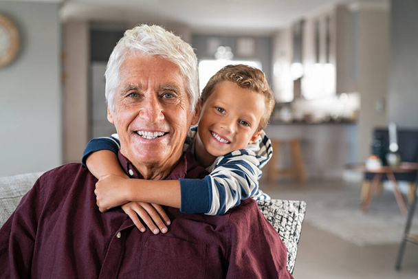 Speelse kleinzoon knuffelt zijn grootvader van achteren terwijl hij thuis naar de camera kijkt. Vrolijk jongetje omarmt oudere man zittend op de bank. Portret van gelukkige oude grootouders glimlachend met zijn kleinkind knuffelen hem van achteren. - Foto, afbeelding