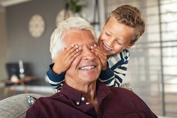 Happy little boy hiding his grandfather eyes with his hands while playing at home. Happy grandson playing hide and seek with senior man at home. Playful child covering eyes of his grandfather with grey hair, guess who game. - 写真・画像