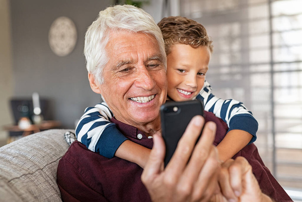 Cute boy hugging old grandfather from behind while sitting on couch and using smartphone at home. Playful grandson embracing senior man while using phone in living room. Elderly grandpa with child watching video on mobile device. - Valokuva, kuva