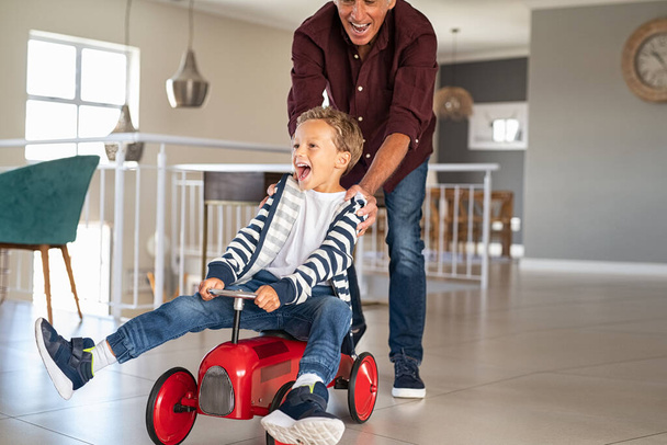 Senior man helping boy ride his toy car at home. Grandfather pushing kid car with little grandson inside to go faster. Excited boy and palyful elderly man playing together at home, copy space. - Photo, image