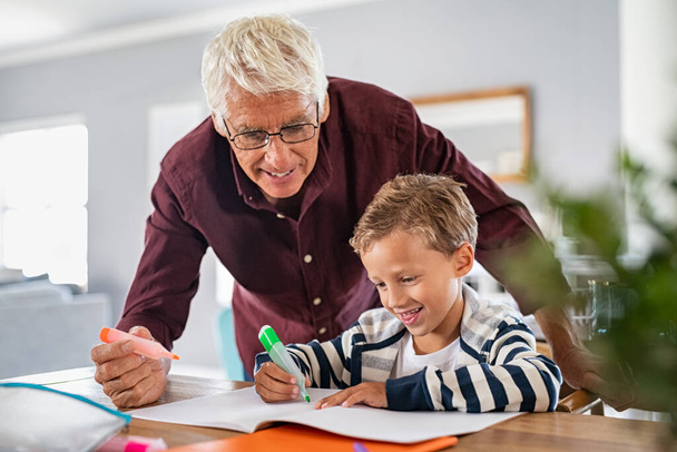Senior man wearing spectacles helping smart boy complete his homework using highlighter pen. Cheerful school boy preparing project in book with grandfather. Grandson and grandpa drawing together on notebook with sketchpen and smiling. - Foto, immagini