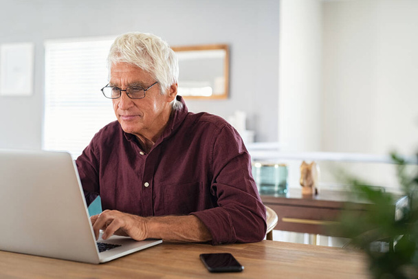 Old man working on laptop at home. Senior man using computer in living room, sitting on chair and looking at screen. Elderly grandfather wearing eyeglasses and working on laptop remotely. - Foto, imagen