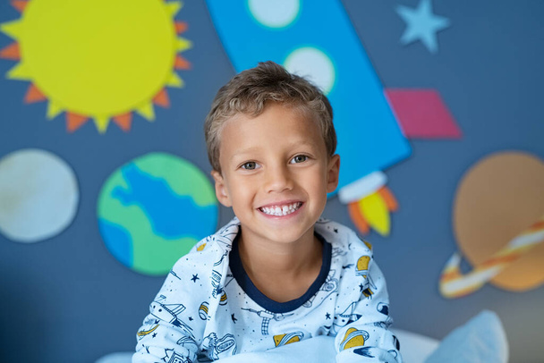 Portrait of smiling boy sitting in space decorated bedroom with the solar system and planets made of cardboard attached to the wall. Portrait of happy cute boy ready to sleep in room with wall decorated with stars and rocket. Joyful kid wearing an as - Foto, Imagem