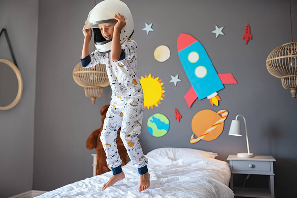 Kleiner süßer Junge im Kosmonautenkostüm, der mit Rakete und Planeten an der Wand auf dem Bett springt. Kind mit Astronautenhelm spielt und träumt davon, Raumfahrer zu werden. Fröhliches Kind trägt Weltraum-Pyjama und hat Spaß mit Sonnensystem und Planeten - Foto, Bild