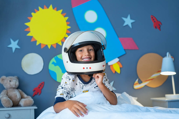 Portrait of happy child wearing astronaut helmet in outer space decorated bedroom. Smiling kid ready to sleep with wall decorated with cardboard planets and rocket. Excited cute boy pretending to be an astronaut looking at camera while sitting on bed - 写真・画像