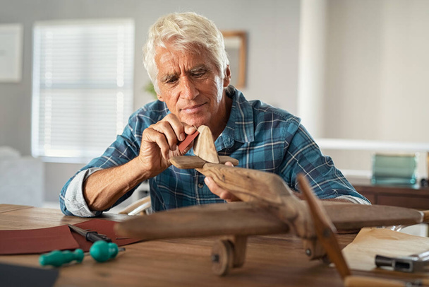 Focused senior man using sandpaper to polish wooden airplane sculpture. Mature carpenter working on making wooden flight at home. Happy old grandfather at home busy making handmade sculpture. - Zdjęcie, obraz
