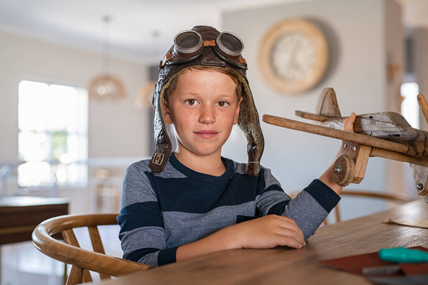 Portrait of little boy wearing pilot helmet playing with wooden airplane at home. School kid playing with wooden plane while looking at camera. Cute child holding wooden handmade airplane model with aviator helmet at home. - Photo, image