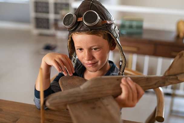 Boy wearing pilot helmet playing with vintage wooden airplane at home. Happy child playing with wooden flight simulator. Little boy holding handmade plane with pilot cap and goggles at home. - Фото, зображення