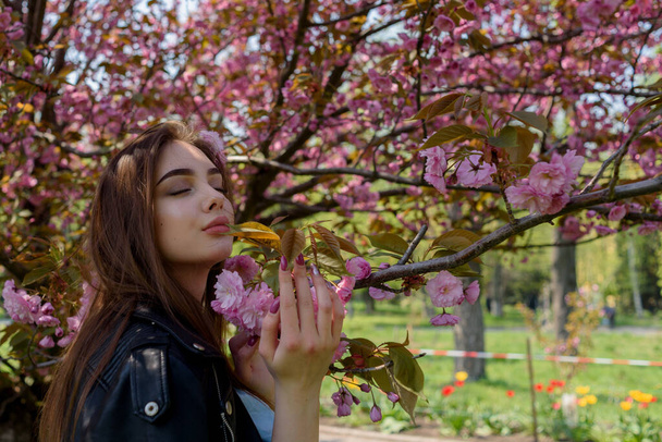 Jeune belle fille aux cheveux longs jouit de la beauté de la nature printanière près de l'arbre sakura en fleurs. Fille dans une veste en cuir dans sakura. - Photo, image