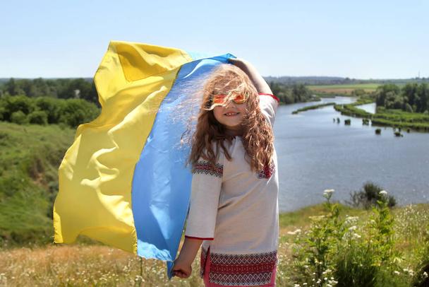 Ευτυχισμένο χαμογελαστό κορίτσι από την Ουκρανία στη Vyshyvanka με κίτρινη και μπλε σημαία σε ένα βουνό στο βάθος των αγρών και ενός ποταμού. Ημέρα Ανεξαρτησίας της Ουκρανίας. Εθνική Σημαία της Ουκρανίας.  - Φωτογραφία, εικόνα
