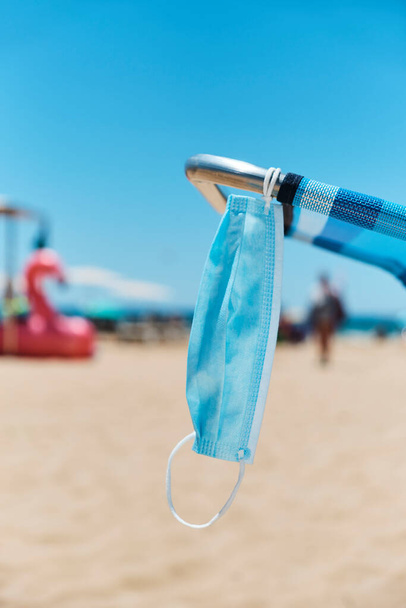 Nahaufnahme einer chirurgischen Maske, die auf einem blauen Liegestuhl am Strand hängt, mit dem Meer im Hintergrund - Foto, Bild