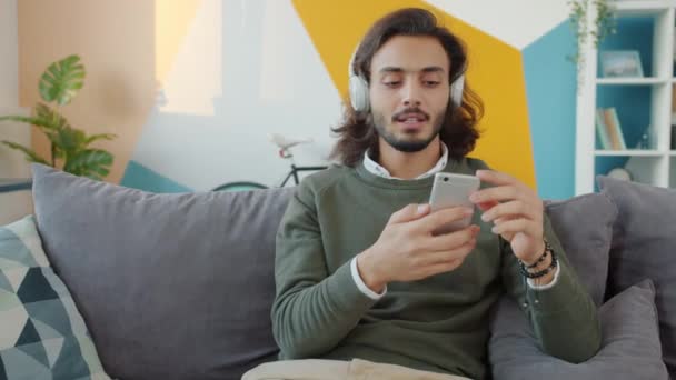 Happy mixed race guy in headphones singing and dancing using smartphone at home - Footage, Video