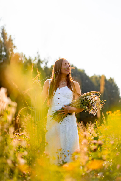 young beautiful woman wit long hair in a white dress and hat among the yellow field of rye and wheat. rustic style concept with long hair in a white sundress in a flowering field - Photo, Image