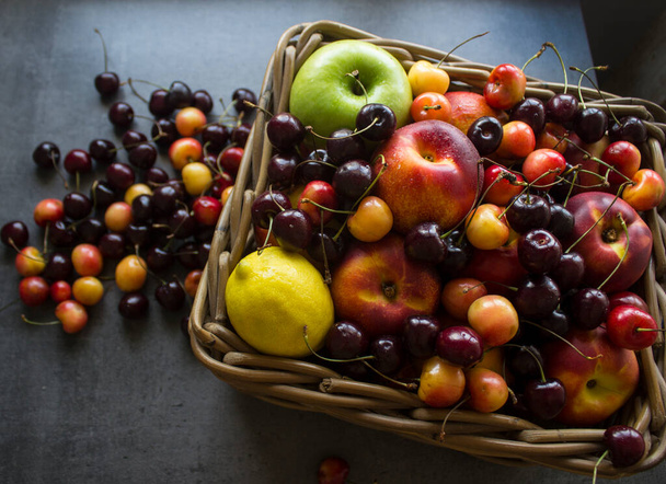 ジューシーな夏の果物や果実でいっぱいのバスケット:チェリー、リンゴ、ネクタリン、マンゴー、レモン。コピースペースとグレーのテクスチャの背景。健康的な食事のコンセプト.  - 写真・画像