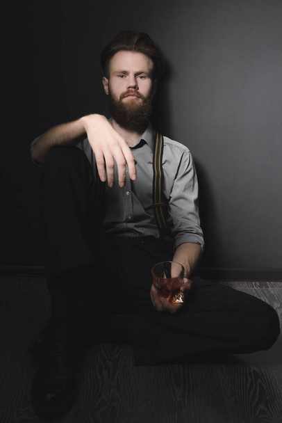 A handsome young man with a beard is sitting in a dark room with a glass of alcohol in his hands. The concept of rest and relaxation, booze and loneliness - Fotoğraf, Görsel