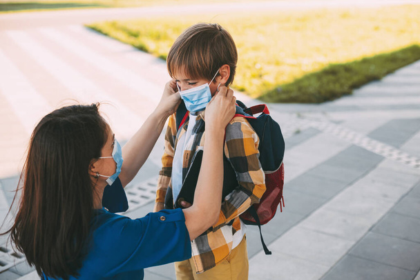 Mama setzt einem Kind eine Maske auf und begleitet es zur Schule oder in den Kindergarten. Ein Junge mit einem maskierten Rucksack während eines Coronavirus. - Foto, Bild
