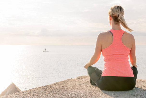back of an unrecognizable blonde woman practicing yoga at sunrise sitting in the lotus position in front of the sea, concept of mental health and relaxation, copy space for text - Photo, image