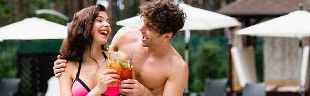 panoramic shot of happy couple clinking cocktails and looking at each other - Фото, изображение
